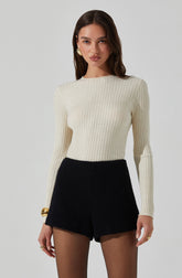 A woman is wearing a long-sleeved, ribbed, cream-colored sweater paired with high-waisted black shorts. She has gold earrings, a large gold bracelet, and rings on her fingers.